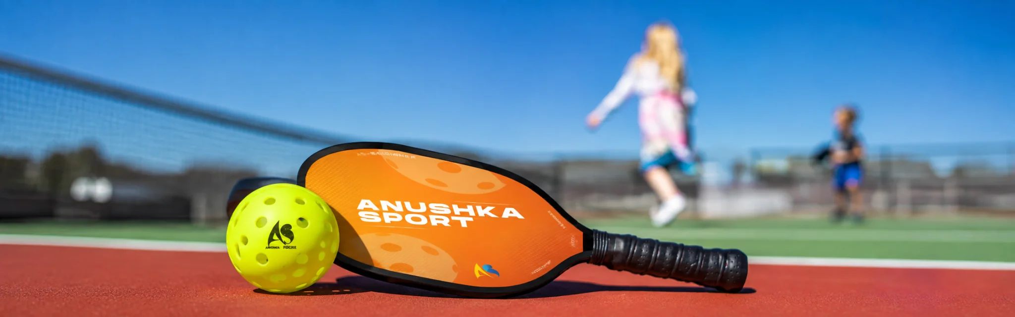 Anushka Sports pickleball paddle and outdoor pickleball ball placed on a pickleball court with players practicing in the background.